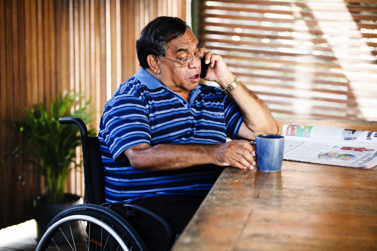 The image shows a man in a wheelchair next to a table. He is looking at a newspaper placed on the table. The man is also talking on the phone and is also holding a mug in his right hand. 