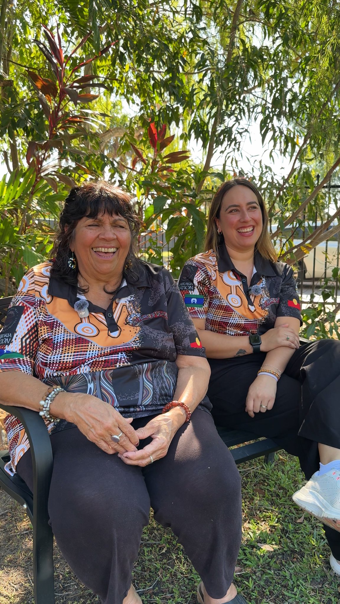 Two women sitting outside on a bench, smiling towards the camera.