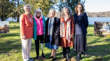 Minister Plibersek, Member for Canberra, Alicia Payne and three National Memorial Advisory Group members stand together outdoors beside a lake during the National Memorial for Victims and Survivors of Institutional Child Sexual Abuse sod-turning ceremony. Display boards showing artist impressions of the future memorial are visible on either side of the group. The ceremony site is surrounded by grass and trees, with water and bushland in the background.