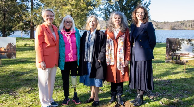 Minister Plibersek, Member for Canberra, Alicia Payne and three National Memorial Advisory Group members stand together outdoors beside a lake during the National Memorial for Victims and Survivors of Institutional Child Sexual Abuse sod-turning ceremony. Display boards showing artist impressions of the future memorial are visible on either side of the group. The ceremony site is surrounded by grass and trees, with water and bushland in the background.