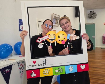 Two people hold a large social media-style frame and emoji props, posing for a photo in a community event setting.