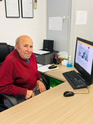 A person sits at a desk using a desktop computer. The Be Connected website is shown on the screen.
